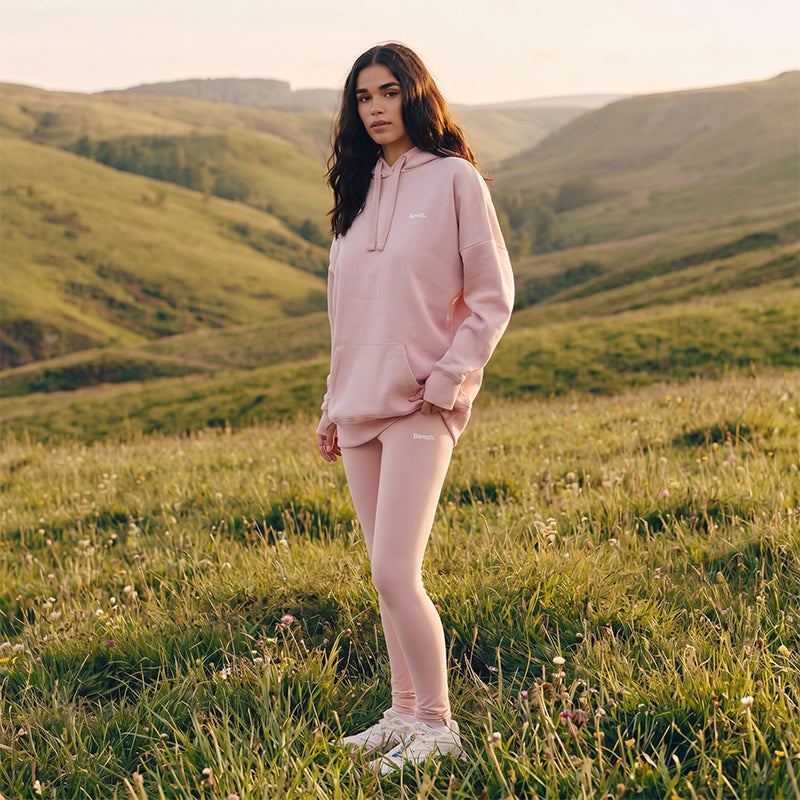 Person wearing a pink hoodie and leggings standing in a grassy field with hills in the background