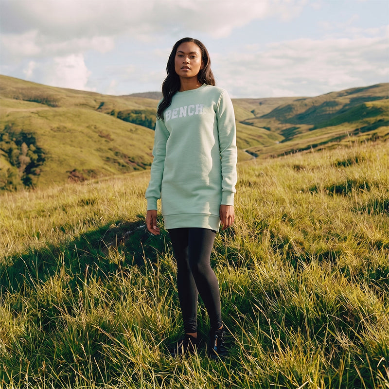 Person wearing a light green sweatshirt with 'BENCH' logo in a grassy field with hills in the background
